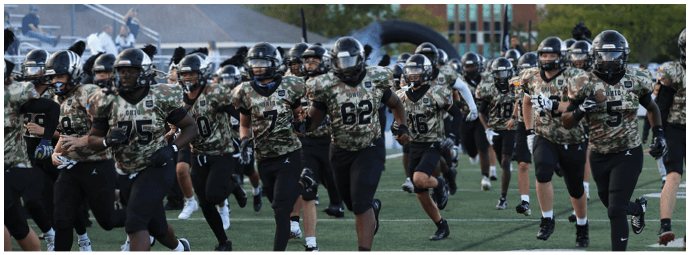A group of people wearing camouflage sports uniforms and helmets are running together on a grassy field, likely participating in a football practice or game. Descripci n generada con IA