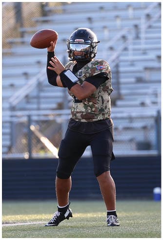 A person wearing a camouflage sports jersey and helmet is poised to throw a football on a field with empty bleachers in the background. Descripci n generada con IA