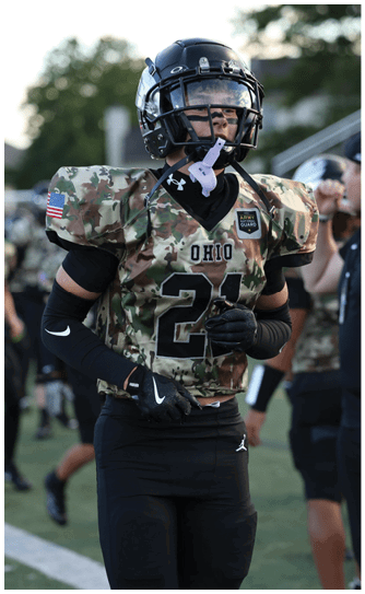 A person wearing a camouflage football uniform with the word Ohio and the number 21 stands on a field with other similarly dressed players in the background. Descripci n generada con IA