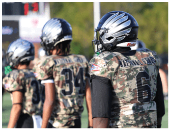 People wearing camouflage football uniforms and helmets stand on a field with one person facing away showing the words “NATIONAL GUARD" and the number 6 on their jersey. Descripci n generada con IA