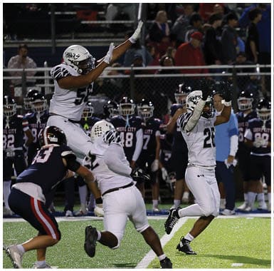 A person wearing a white football uniform and helmet raises both hands while another person in a dark uniform stands in the background. Descripci n generada con IA