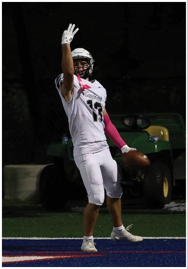 A person in a white football uniform is standing in the end zone holding a football and raising three fingers. Descripci n generada con IA