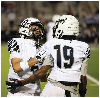 Two people wearing white football helmets and uniforms appear to be engaged in a conversation or interaction on a field. Descripci n generada con IA
