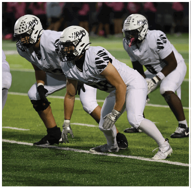 A person in white football gear with a helmet labeled “HAWKS" is crouched in a ready position on a football field with another similarly dressed person in the background. Descripci n generada con IA