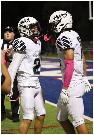 Two people wearing football helmets and white jerseys with pink accessories are facing each other on a sports field. Descripci n generada con IA