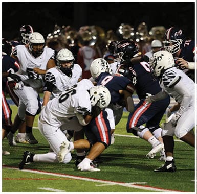 Players in white and dark uniforms are engaged in a football game on a field with a marching band visible in the background. Descripci n generada con IA