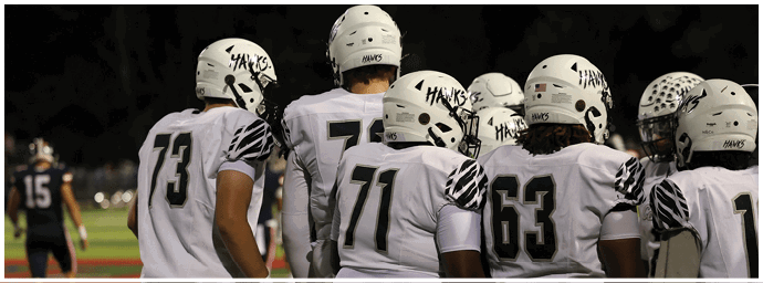 A group of people wearing white football uniforms and helmets with the word “Hawks" on them are standing together on a field. Descripci n generada con IA