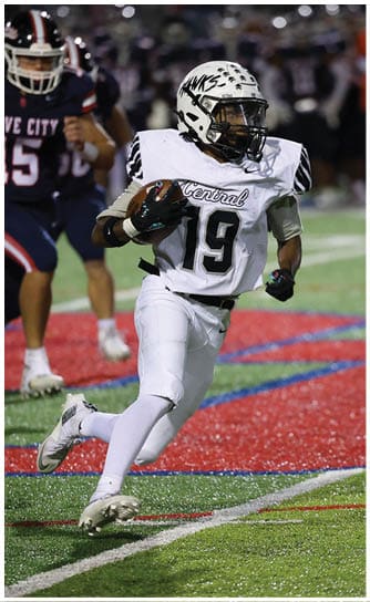 A person wearing a white football uniform with the number 19 is running on a field with a red and blue logo while holding a football. Descripci n generada con IA