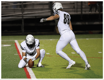 A person in white football gear is kneeling on a field holding a football upright on the ground while another person in similar attire stands nearby. Descripci n generada con IA