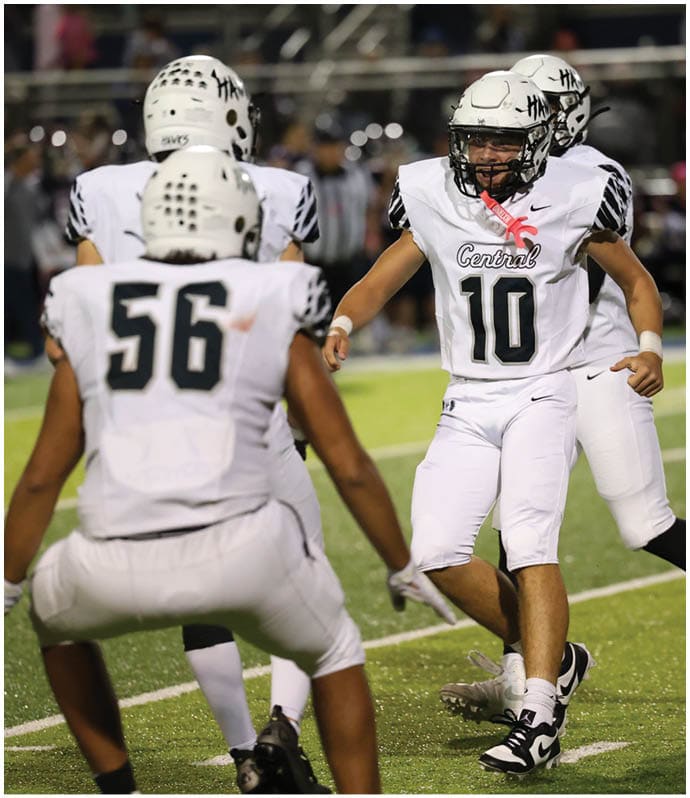 A group of people wearing white football uniforms with helmets are on a field, with one person in the center appearing to celebrate or communicate with teammates. Descripci n generada con IA