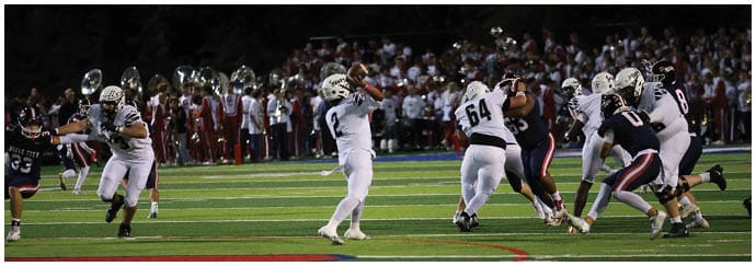 A group of football players in white and dark uniforms are engaged in a play on a well-lit field with a crowd in the background. Descripci n generada con IA