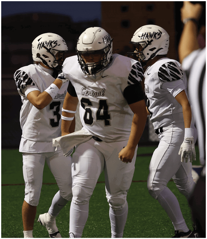 Three people in white football uniforms with “Hawks" helmets are on a field at dusk, with one person in the center wearing number 84 and the others nearby, while a referee raises an arm in the foreground. Descripci n generada con IA
