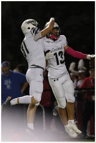 Two football players in white uniforms celebrate on the field at night while a referee and spectators watch in the background. Descripci n generada con IA