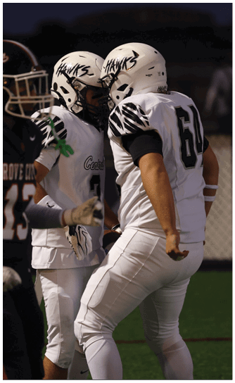 Two people in white football uniforms and helmets with “HAWKS" written on them are facing each other closely on a field during a game. Descripci n generada con IA