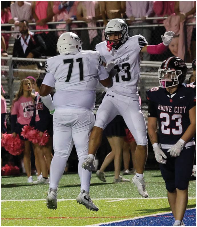 Two football players in white uniforms celebrate near the end zone while a crowd of cheerleaders in pink and spectators watch from the stands. Descripci n generada con IA
