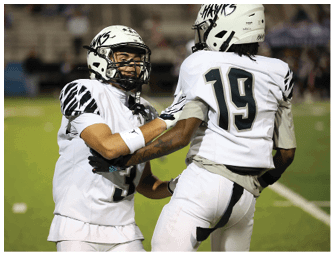 Two people wearing white football uniforms and helmets are engaged in a play on a football field. Descripci n generada con IA