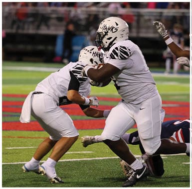 Two football players in white uniforms are engaged in a play while three players in dark uniforms attempt to tackle or block them on a field with a crowd in the background. Descripci n generada con IA