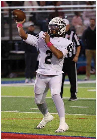 A person wearing a white football uniform with the number 2 is preparing to throw a football on a field with red and blue markings. Descripci n generada con IA