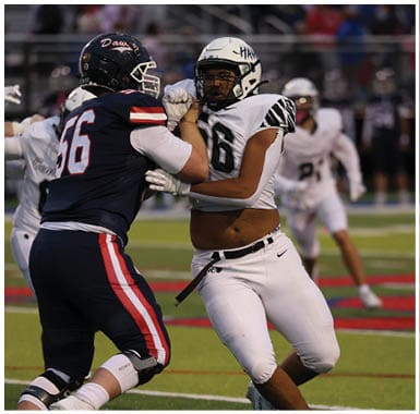 Two people in football uniforms are engaged in a play on a football field with one person in white running while being blocked by a person in a dark uniform. Descripci n generada con IA
