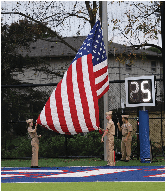 Four people in uniform are raising a large American flag outdoors near a scoreboard displaying the number 25. Descripci n generada con IA