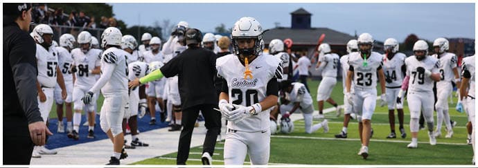 A group of people wearing white football uniforms and helmets are gathered on a sports field with one person in the foreground walking towards the camera. Descripci n generada con IA