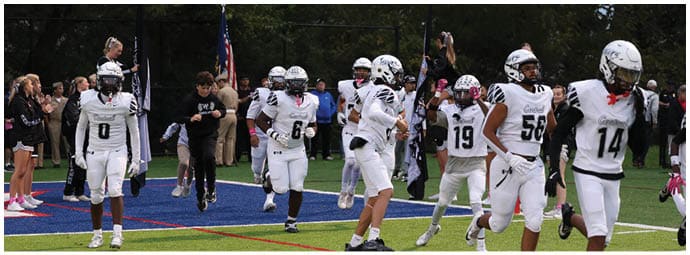 People in white football uniforms are running onto a field while others stand on the sidelines, with some holding flags and a crowd in the background. Descripci n generada con IA