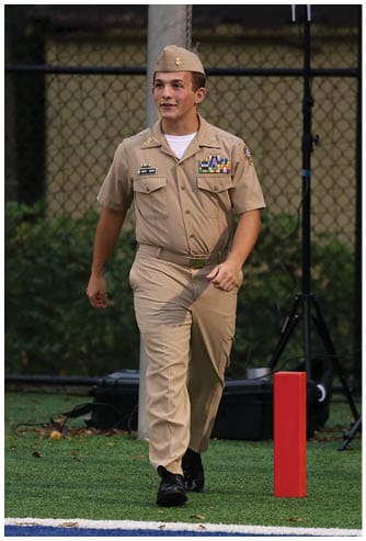 A person in a tan military uniform with a cap is walking on a sports field with green turf and a blue and red painted area. Descripci n generada con IA