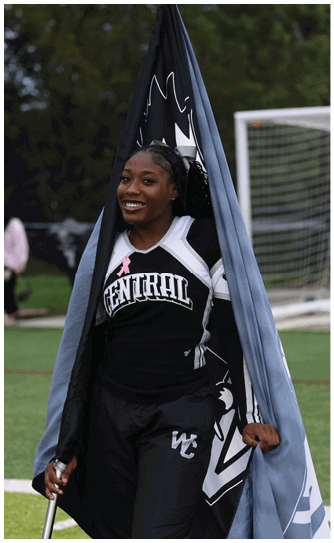 A person wearing a Central cheerleading uniform is smiling and holding a large flag on a sports field with blurred people in the background. Descripci n generada con IA