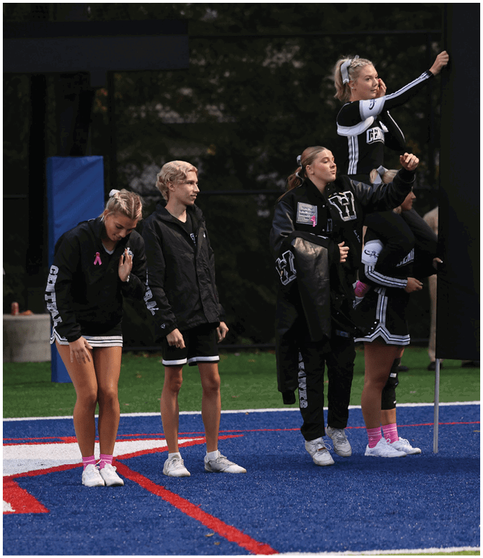 Five people in cheerleading uniforms and jackets stand on a sports field with blue turf, with one person lifted in a cheer pose near a large black banner. Descripci n generada con IA