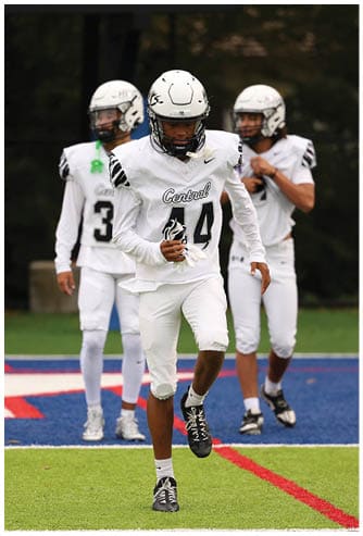 Three people in white football uniforms and helmets are on a field, with one person running toward the camera while the others stand behind. Descripci n generada con IA