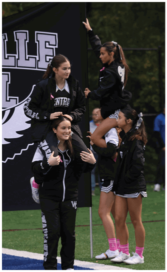Four people in athletic clothing are on a sports field with two individuals sitting on the shoulders of others near a large black and white banner. Descripci n generada con IA