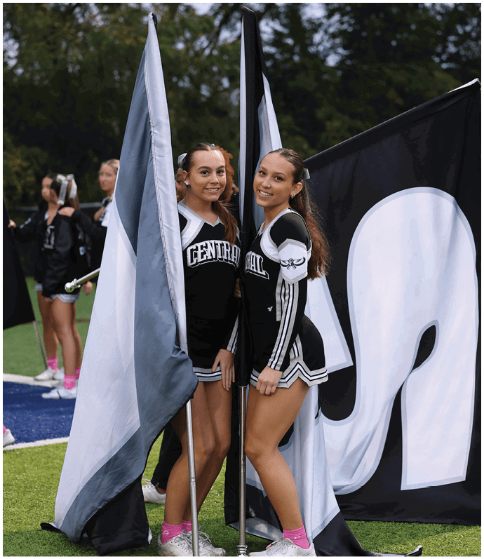 Two people in cheerleading uniforms stand on a sports field holding large flags with a group of similarly dressed people in the background. Descripci n generada con IA