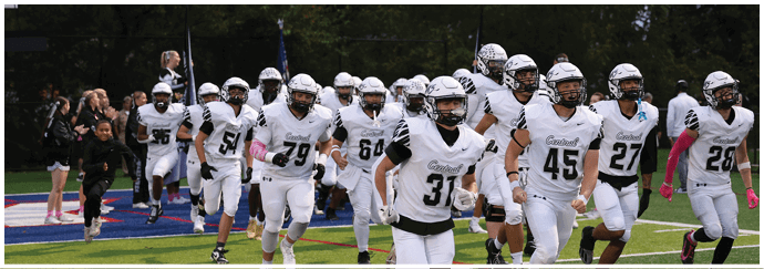 A group of people wearing white football uniforms and helmets are running onto a field with a crowd and cheerleaders in the background. Descripci n generada con IA