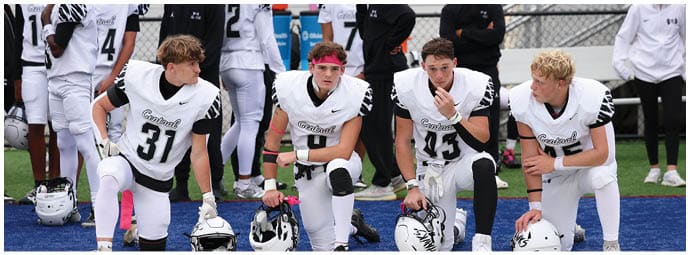 Four people in white football uniforms kneel on a field, appearing to be in conversation or preparing for a game. Descripci n generada con IA