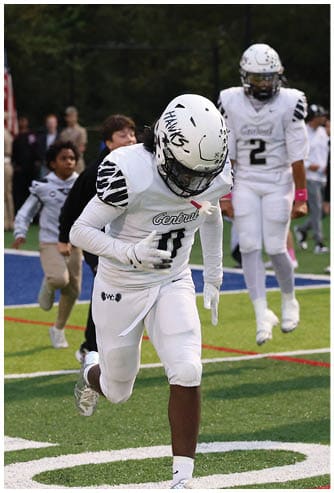 A person in a white football uniform and helmet is running on a field with other people and flags visible in the background. Descripci n generada con IA
