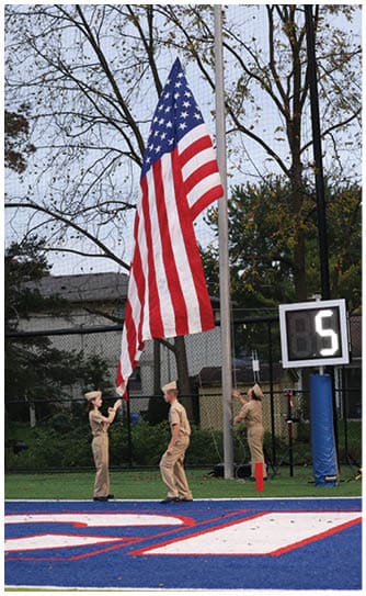 Three people in uniform are raising an American flag on a sports field near a scoreboard displaying the number five. Descripci n generada con IA