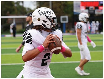 An adult wearing a white football uniform with black stripes and a helmet labeled “HAWKS" is holding a football and preparing to throw it on a green field with other players in the background. Descripci n generada con IA