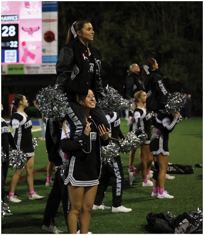 People in cheerleading uniforms with pom-poms perform a routine on a sports field at night with a scoreboard visible in the background. Descripci n generada con IA
