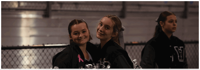 Two people wearing dark jackets with pink ribbons smile and pose closely together outdoors near a chain-link fence. Descripci n generada con IA