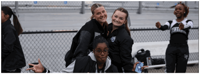 Four people wearing athletic clothing are smiling and posing near a chain-link fence with bleachers in the background. Descripci n generada con IA