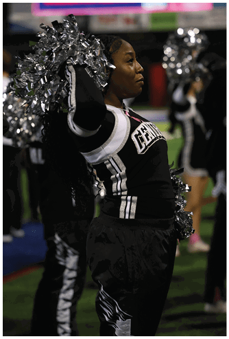 A person in a cheerleading uniform with pom-poms is performing on a field with other cheerleaders visible in the background. Descripci n generada con IA