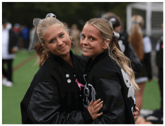 Two adults wearing black letterman jackets with hair tied back are smiling and embracing each other outdoors on a sports field. Descripci n generada con IA