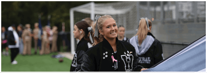 A person wearing a letterman jacket with a pink ribbon pin is smiling outdoors near a sports field with other people in the background. Descripci n generada con IA