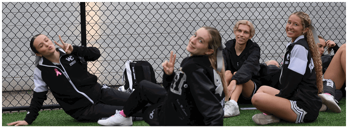 Three people wearing black jackets are sitting near a chain-link fence by a body of water, with two of them making peace signs and smiling. Descripci n generada con IA