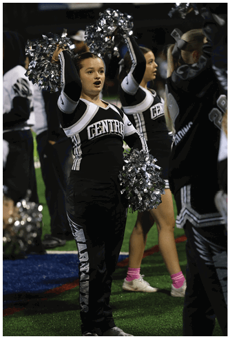 Two adults in black and white cheerleading uniforms with “CENTRAL" on the front are performing with silver pom-poms, one in the foreground with an open mouth and the other slightly blurred in the background. Descripci n generada con IA