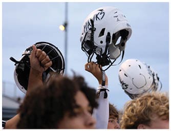 A person is holding up a white football helmet with the word “HAWKS" written on it while other similar helmets are visible in the background against a cloudy sky. Descripci n generada con IA