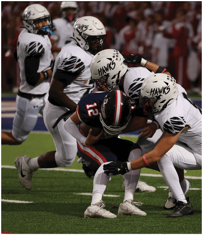 A person in a white football uniform with “HAWKS" written on the helmet is crouched on a green field during a game. Descripci n generada con IA