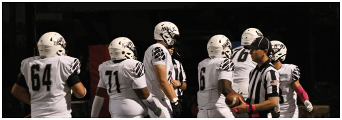 A group of people wearing football helmets and uniforms are gathered on a field under stadium lights with advertising banners visible in the background. Descripci n generada con IA