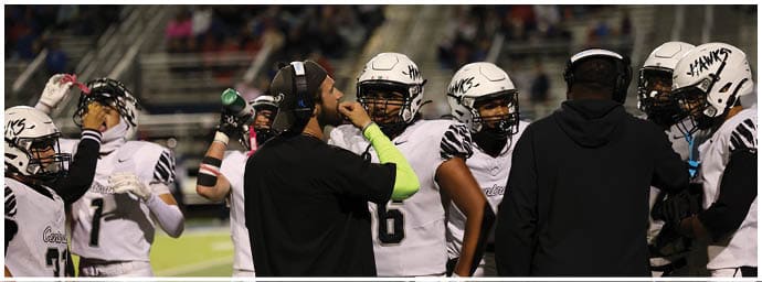 A group of people wearing football helmets and uniforms are gathered on a field with one person in a cap and headset giving instructions. Descripci n generada con IA