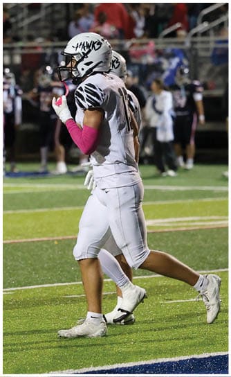 Two people in dark football uniforms with helmets stand on a field facing a person in a white football uniform and helmet, with a crowd visible in the background. Descripci n generada con IA